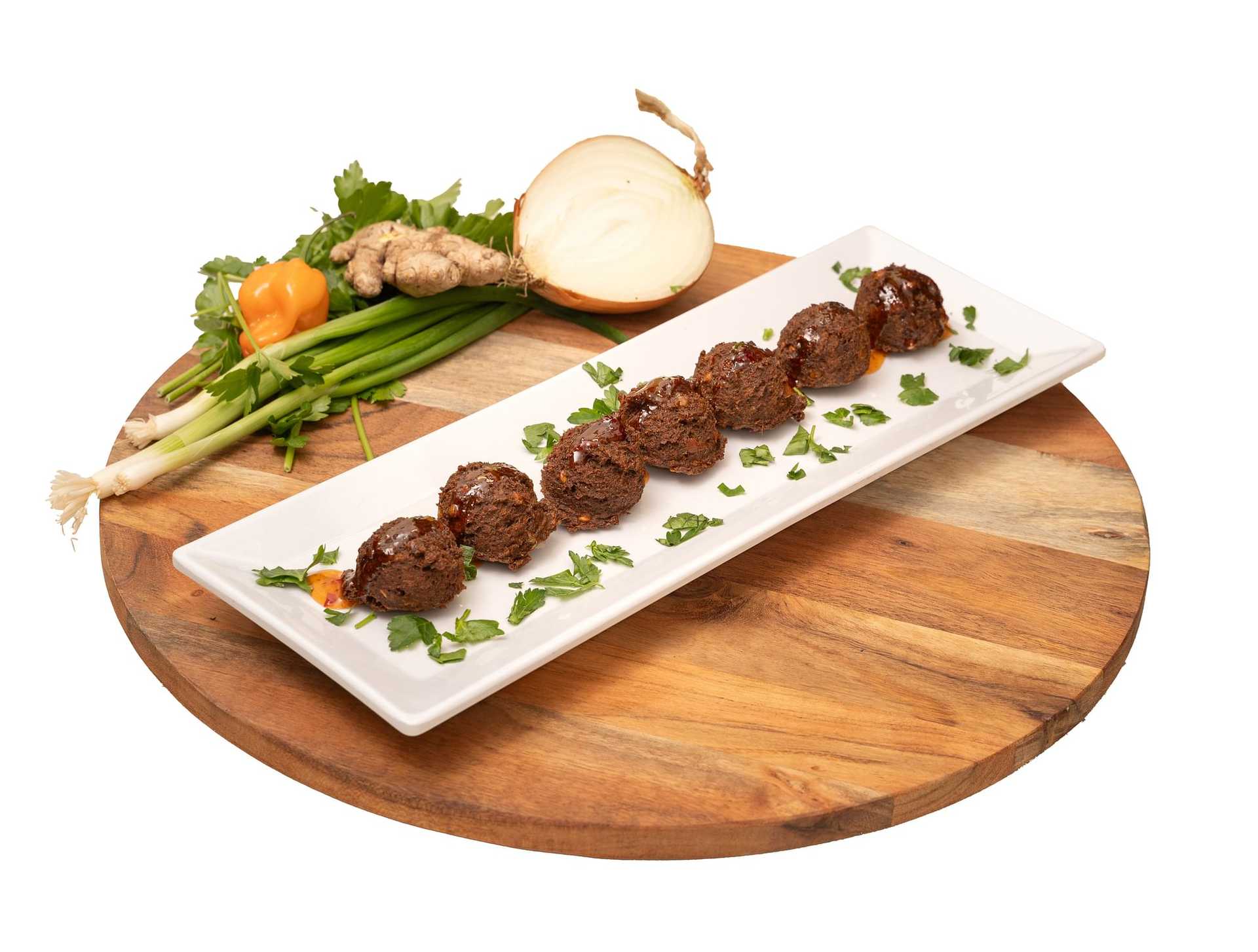 Plate of served meatballs garnished with herbs, surrounded by vegetables and spices on a wooden board.